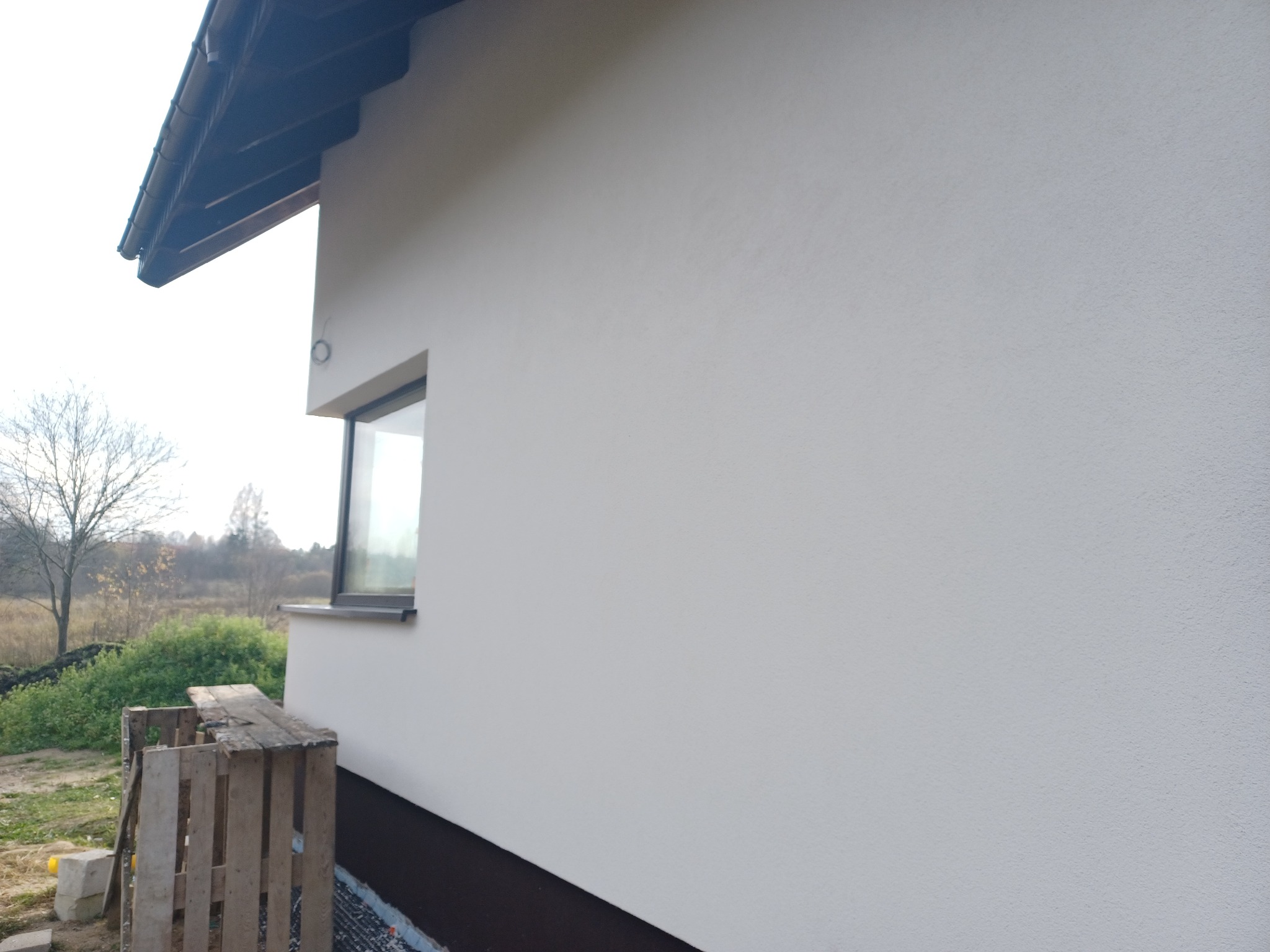 Fragment ściany domu z oknem po wykonaniu elewacji, widoczna faktura tynku, w tle drzewa i krzewy, na pierwszym planie paleta.