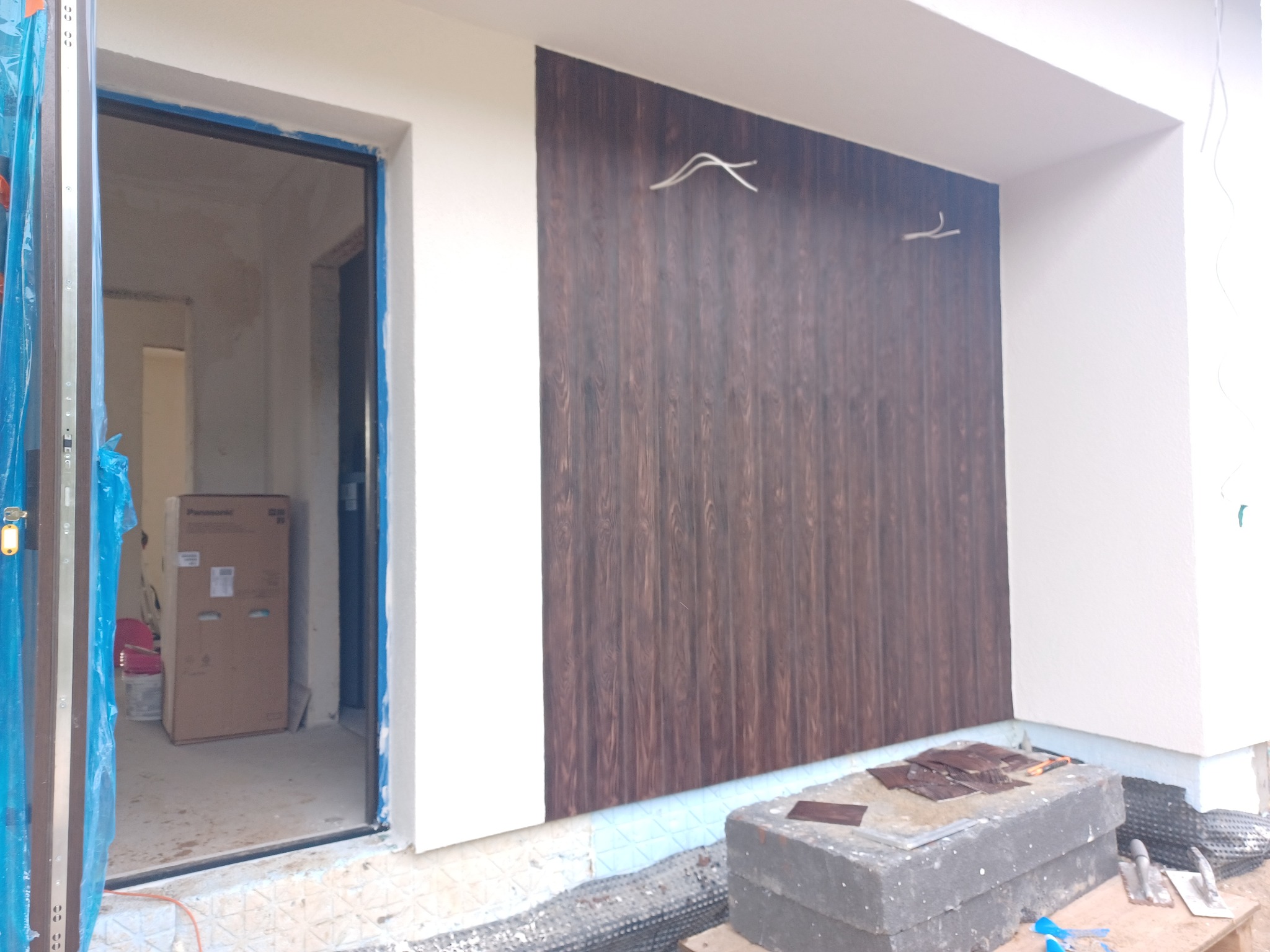 Montaż paneli elewacyjnych imitujących drewno na ocieplonym budynku, widoczne przewody elektryczne wystające ze ściany, przygotowane płytki i narzędzia na bloczkach betonowych, otwarte drzwi...