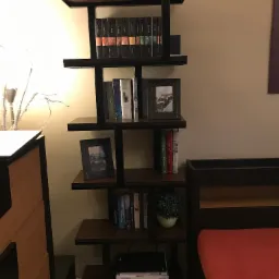 Ręcznie robiony regał na książki z ciemnego drewna i metalu, wypełniony książkami, ramkami ze zdjęciami i małą rośliną doniczkową, ustawiony w sypialni obok łóżka z czerwoną pościelą.