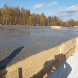 Wylany beton na przygotowany fundament, widoczne drewniane szalunki i białe bloczki fundamentowe, w tle drzewa z jesiennymi liśćmi i błękitne niebo.