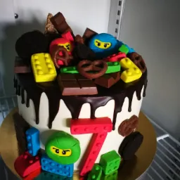 Tort urodzinowy z motywem Lego Ninjago, dekorowany czekoladą, preclami i ciasteczkami Oreo, z cyfrą 7 na froncie, na złotym podkładzie.