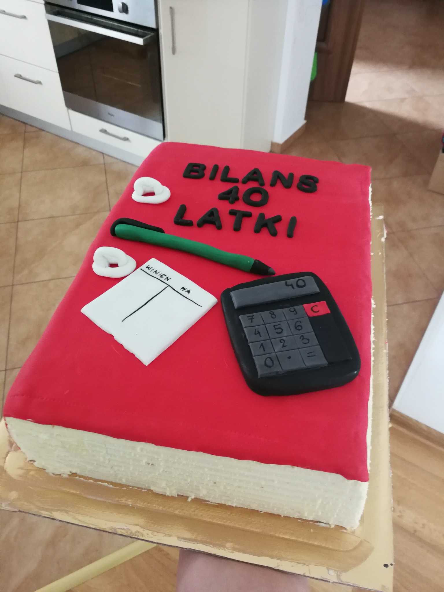 Tort w kształcie czerwonej księgi bilansowej z napisem 'Bilans 40 latki', ozdobiony kalkulatorem, długopisem i kartką z tabelą 'Winien/Ma'.