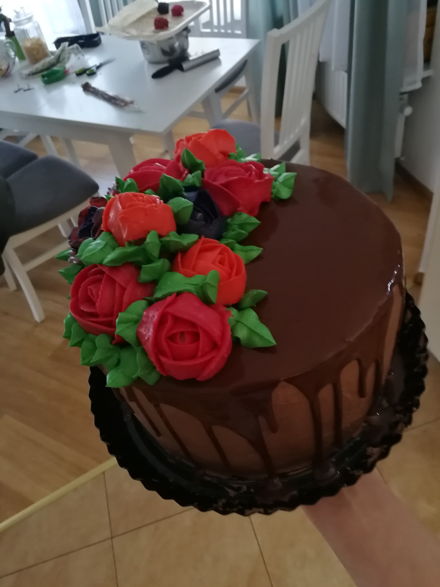 Okrągły tort czekoladowy udekorowany kremowymi różami w odcieniach czerwieni i pomarańczy, z polewą spływającą po bokach, trzymany w dłoni na tle domowego wnętrza z białym stołem i krzesłami.