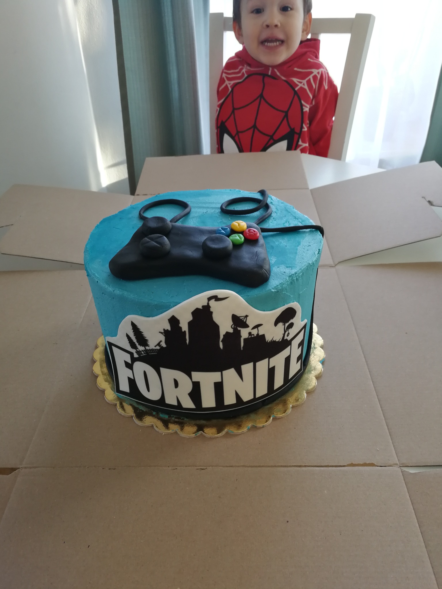 Turkusowy tort z motywem Fortnite i kontrolerem do gier, na złotym podkładzie, w tle uśmiechnięty chłopiec w bluzie Spidermana.