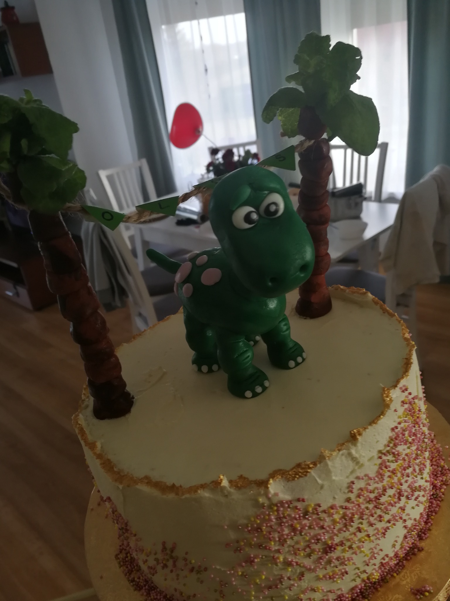 Dwupoziomowy tort urodzinowy z kremem, posypką i figurką zielonego dinozaura oraz palmami z liści na górze, dekorowany girlandą z napisem.
