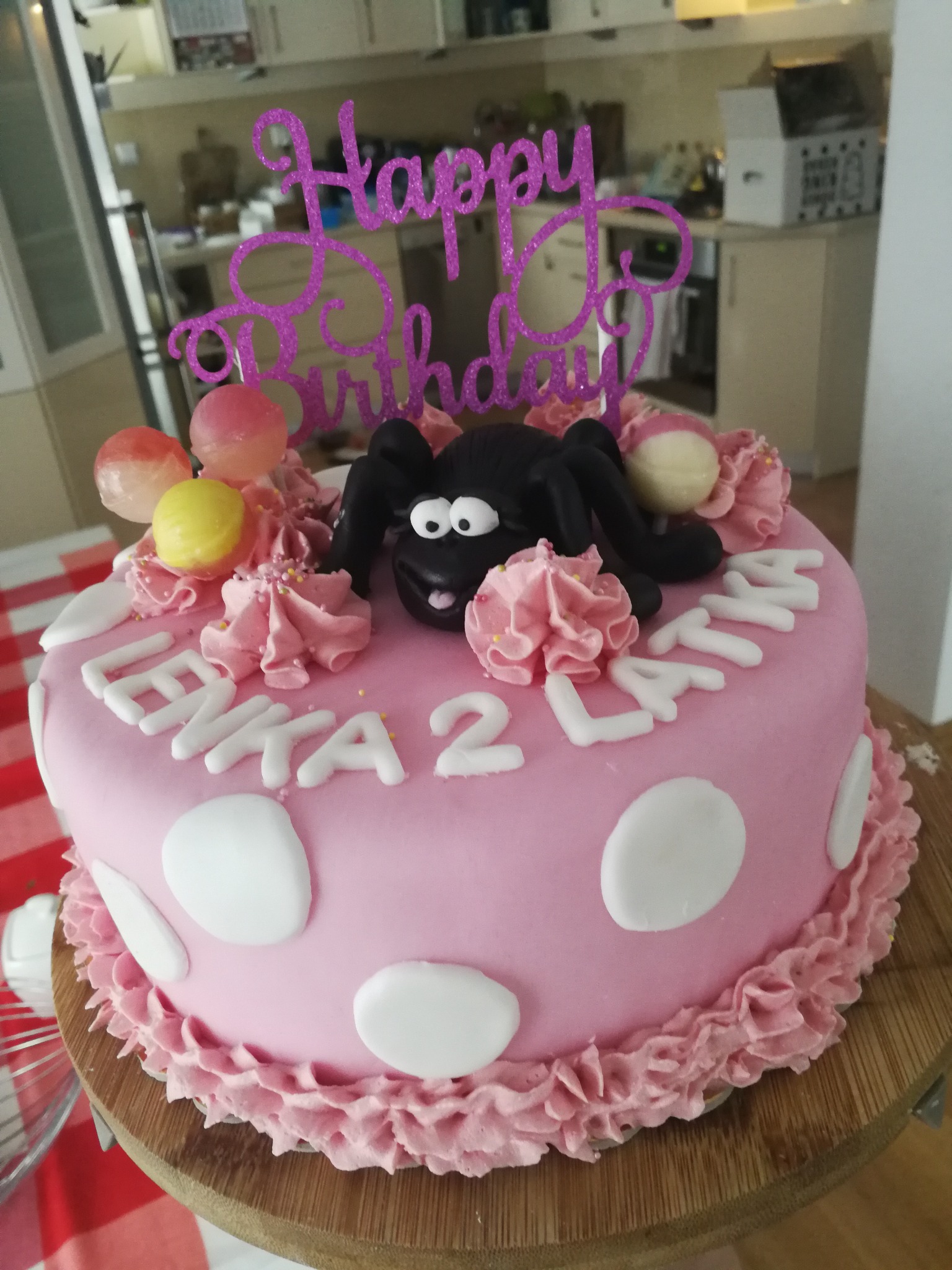 Różowy tort urodzinowy z napisem 'Lenka 2 Latka', dekorowany lizakami, rozetkami z kremu i czarną figurką owieczki Shaun, zwieńczony brokatowym napisem 'Happy Birthday'.
