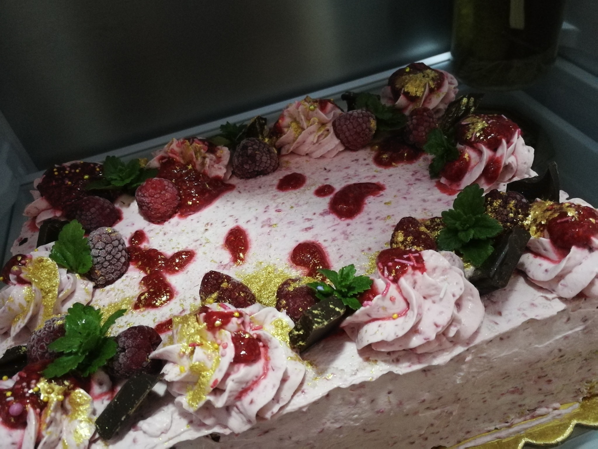 Prostokątny tort z kremem malinowym, świeżymi malinami, kawałkami czekolady, listkami mięty i złotymi płatkami, widoczne krople dżemu malinowego.