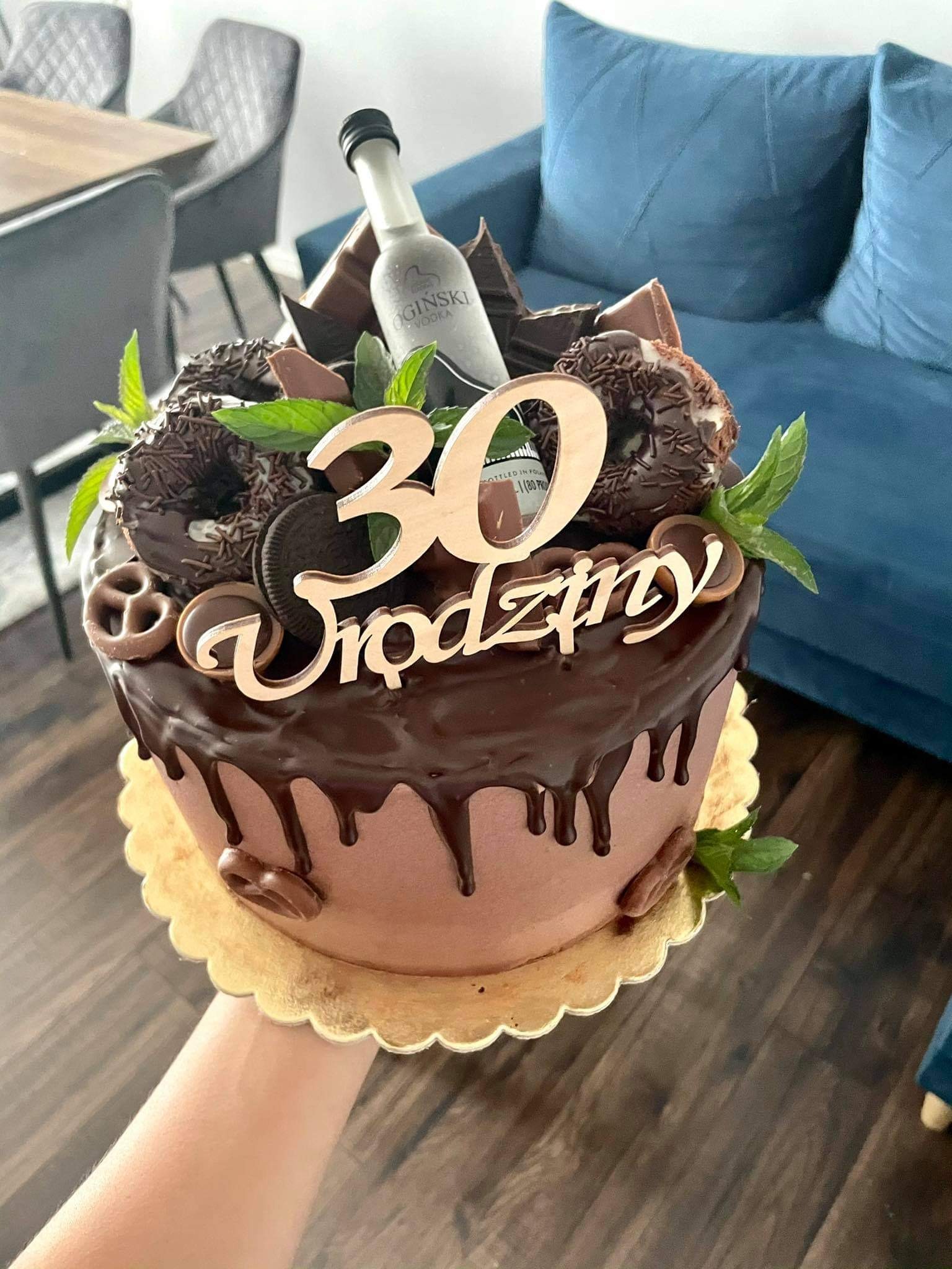 Tort urodzinowy z czekoladowym ganache, dekorowany pączkami, preclami, miętą i butelką wódki na 30 urodziny, trzymany w dłoni.