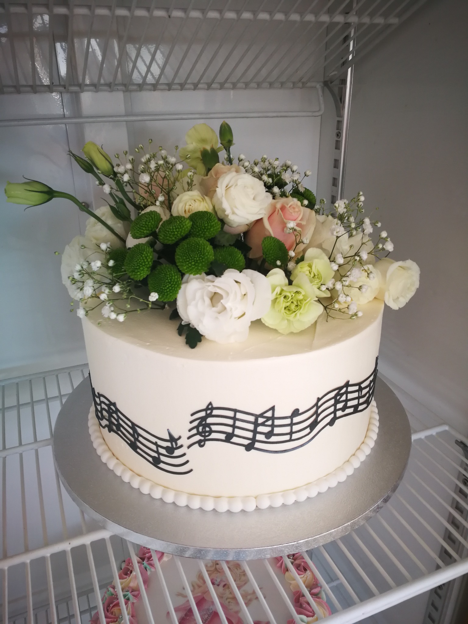 Tort z motywem muzycznym i dekoracją z białych i zielonych kwiatów, umieszczony na srebrnej paterze w chłodnej przestrzeni.