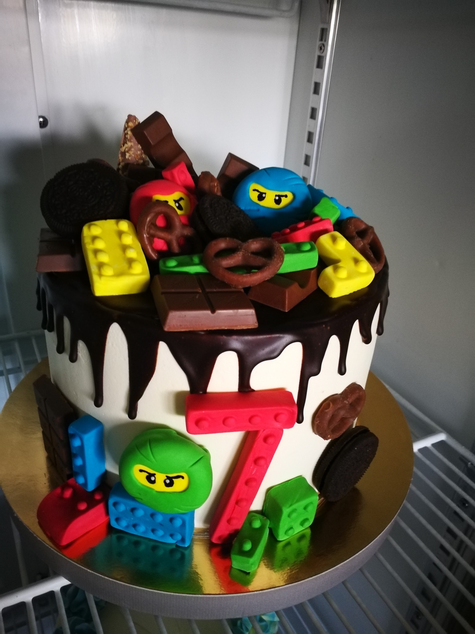 Tort urodzinowy z motywem Lego Ninjago, dekorowany czekoladą, preclami i ciasteczkami Oreo, z cyfrą 7 na froncie, na złotym podkładzie.