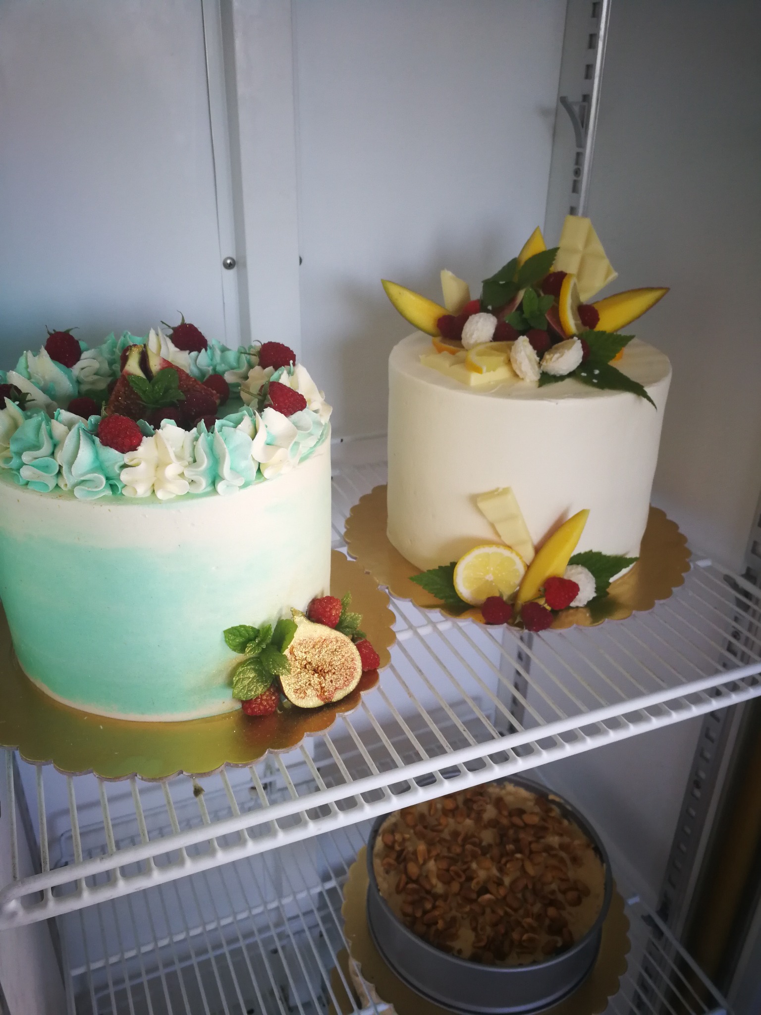 Dwa torty na złotych podkładkach, jeden z kremem w kolorze ombre turkusowo-białym i malinami, drugi biały z dekoracją z mango, cytryny i białej czekolady, na półce lodówki, na dole widoczny tort...