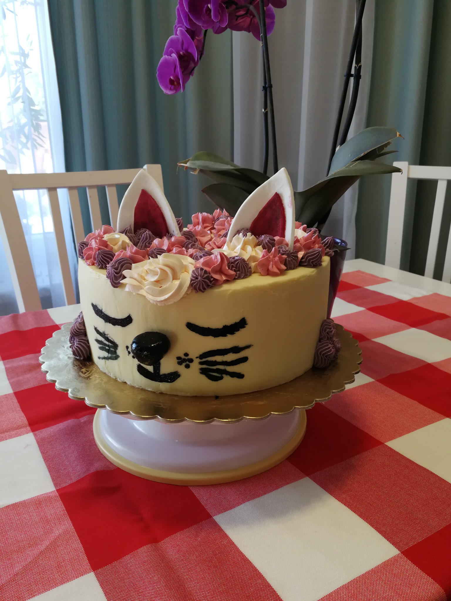 Tort w kształcie kota z uszami z masy cukrowej, kremowymi dekoracjami i czarnymi detalami na pyszczku, umieszczony na złotej paterze na stole z czerwoną kratą, w tle storczyk.