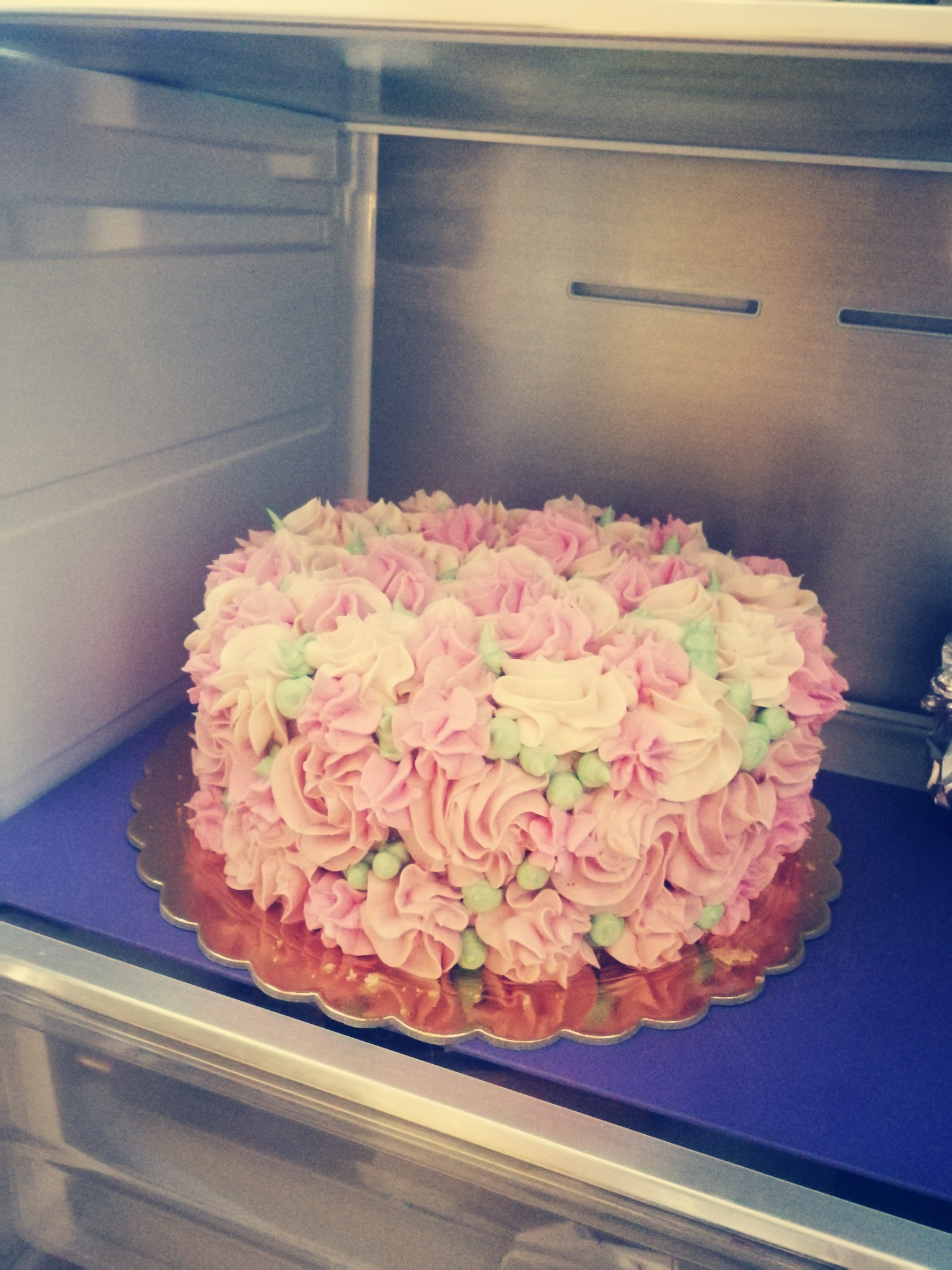 Tort zdobiony kremem w odcieniach różu i zieleni, ułożony w lodówce na złotym podkładzie i fioletowej półce.