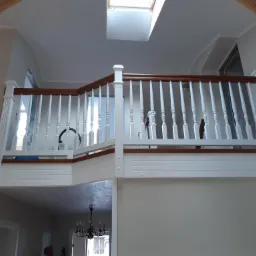 Biała barierka z drewnianą poręczą na antresoli, widok z dołu na sufit z oknem dachowym, widoczne elementy konstrukcji drewnianej dachu.