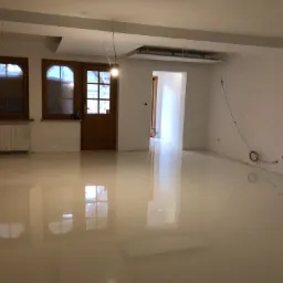 Błyszcząca, beżowa posadzka żywiczna w jasnym pomieszczeniu w Lipnicy Małej, z widocznymi odbiciami okien i drzwi, oraz wystającymi przewodami elektrycznymi ze ściany.