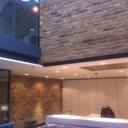 Usługi Wykończeniowe - Wnętrze z ceglaną ścianą, nowoczesnym oświetleniem i minimalistycznymi białymi szafkami, widoczny fragment szklanej ściany z odblaskami.