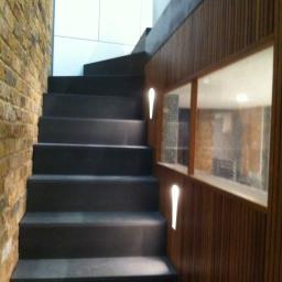 Usługi Wykończeniowe - Nowoczesne schody z ciemnymi stopniami przylegające do ściany z cegły i drewnianej boazerii z oknem podświetlonym wąskimi lampami.