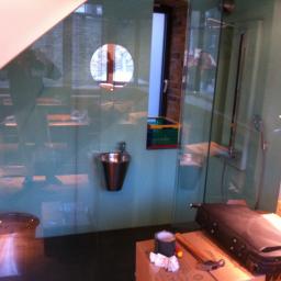 Usługi Wykończeniowe - Niewielka, metalowa umywalka zamontowana na ścianie w surowym pomieszczeniu, widoczne narzędzia i akcesoria na kartonowym pudle, odbicie osoby robiącej zdjęcie.