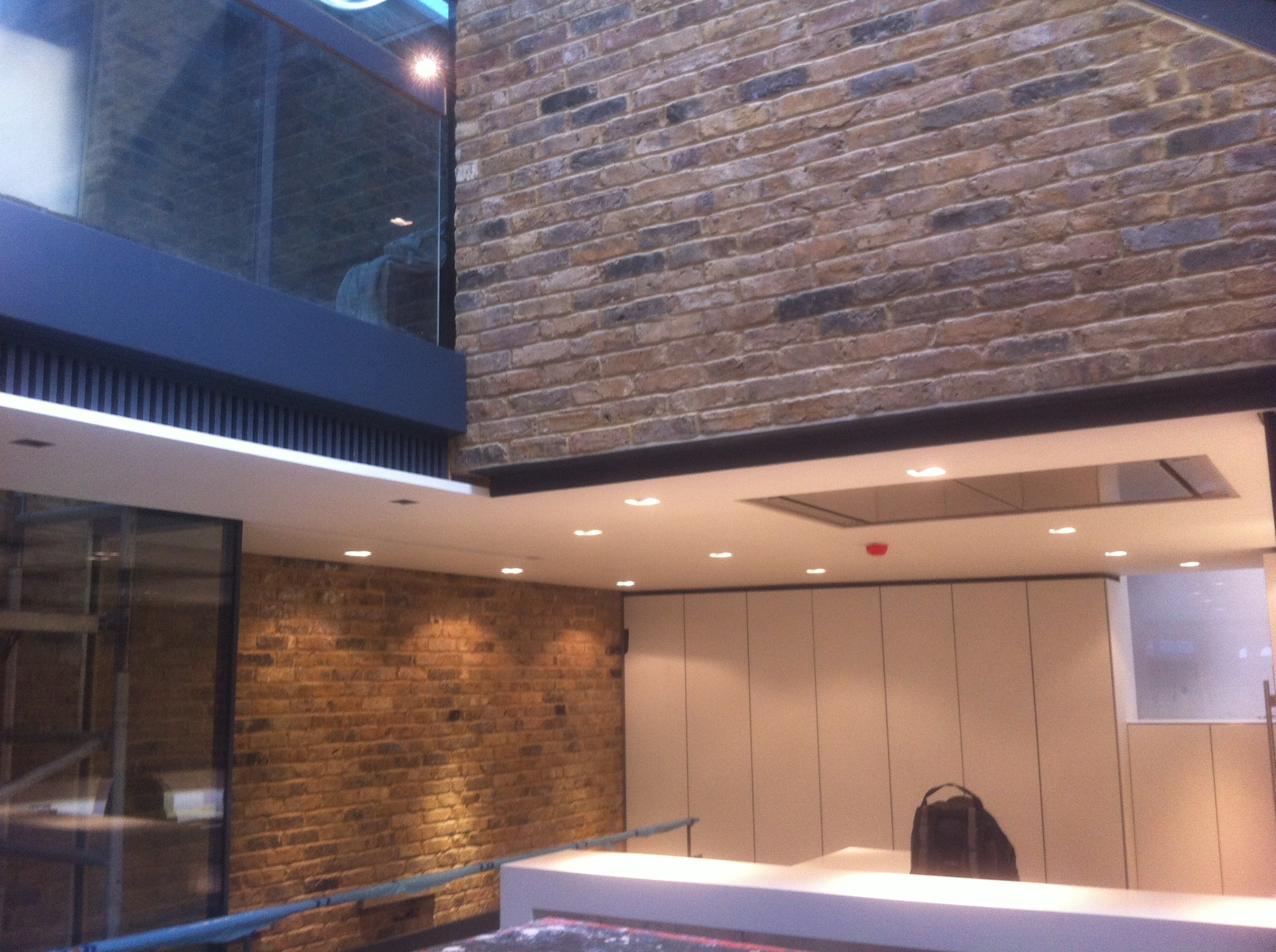 Wnętrze z ceglaną ścianą, nowoczesnym oświetleniem i minimalistycznymi białymi szafkami, widoczny fragment szklanej ściany z odblaskami.