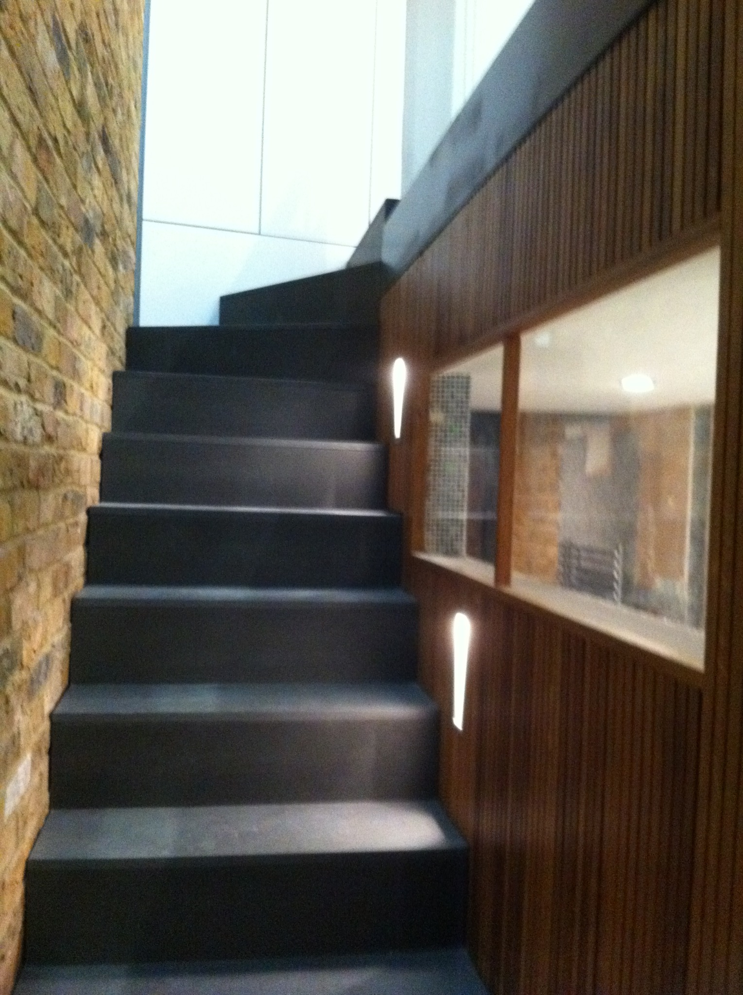 Nowoczesne schody z ciemnymi stopniami przylegające do ściany z cegły i drewnianej boazerii z oknem podświetlonym wąskimi lampami.