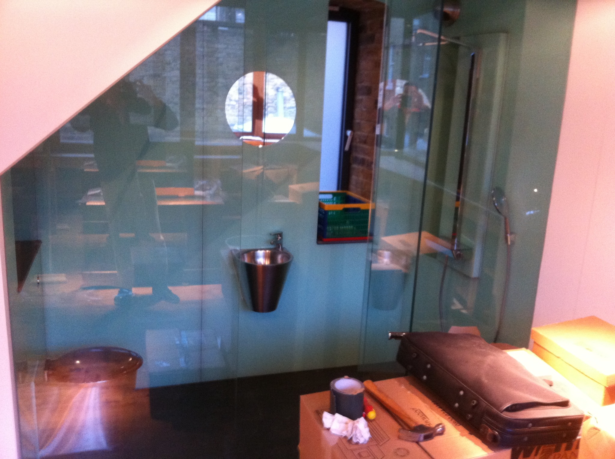 Niewielka, metalowa umywalka zamontowana na ścianie w surowym pomieszczeniu, widoczne narzędzia i akcesoria na kartonowym pudle, odbicie osoby robiącej zdjęcie.