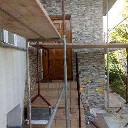 Rem-bud - Elewacja budynku w trakcie wykończenia z użyciem szarej, kamiennej okładziny, widoczne rusztowanie, schody wejściowe i częściowo zamontowane drzwi wejściowe.