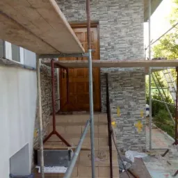 Elewacja budynku w trakcie wykończenia z użyciem szarej, kamiennej okładziny, widoczne rusztowanie, schody wejściowe i częściowo zamontowane drzwi wejściowe.