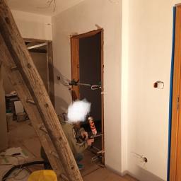 Rem-bud - Montaż ościeżnicy w Tarnowie: fachowiec uszczelnia przestrzeń między ścianą a drewnianą ramą drzwi przy użyciu pistoletu do pianki montażowej, widoczna drabina i odkurzacz przemysłowy.