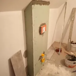 Stelaż podtynkowy obudowany płytą karton gips, z widocznymi przyłączami wodnymi i puszką instalacyjną JOMO, w trakcie prac wykończeniowych łazienki.