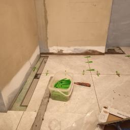 Rem-bud - Fragment łazienki w trakcie prac wykończeniowych: ułożone płytki podłogowe z zielonymi krzyżykami dystansowymi, widoczny pojemnik z zapasem krzyżyków, nóż tapicerski i paczka płytek z napisem 'MADE...