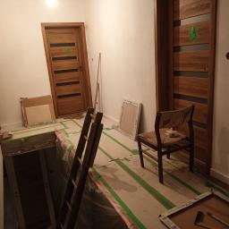 Rem-bud - Wnętrze w trakcie prac wykończeniowych: widoczne zabezpieczone podłoże, drabina, krzesło, oraz zamontowane drewniane drzwi z zieloną taśmą zabezpieczającą, obok drzwi w trakcie montażu.