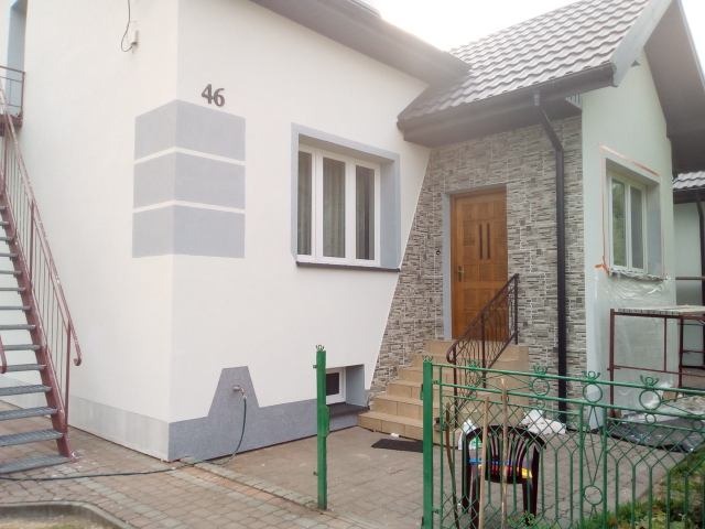 Wykończona elewacja domu jednorodzinnego z numerem 46, z drewnianymi drzwiami wejściowymi i schodami z metalową barierką, fragment elewacji wykończony imitacją kamienia, pozostała część w kolorze...