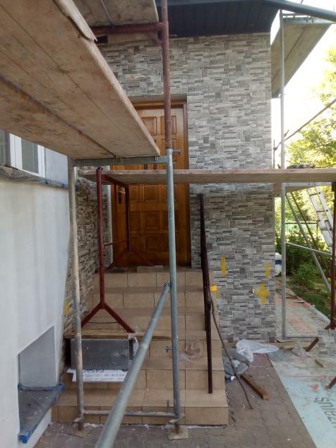 Elewacja budynku w trakcie wykończenia z użyciem szarej, kamiennej okładziny, widoczne rusztowanie, schody wejściowe i częściowo zamontowane drzwi wejściowe.