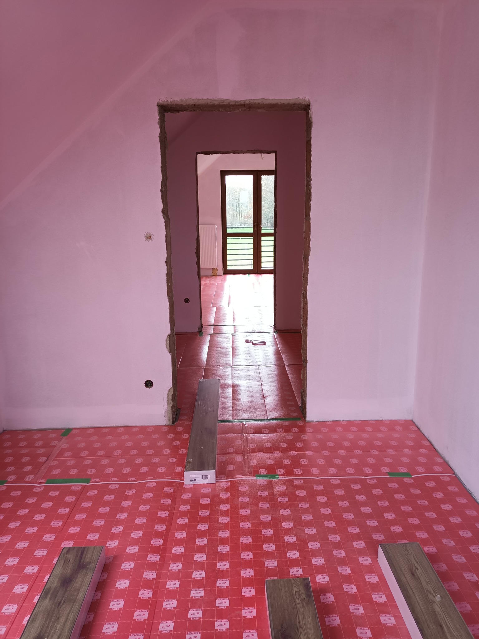 Przygotowanie do ułożenia paneli: widoczna podkładka pod panele w kolorze różowym z nadrukowanymi napisami oraz kilka paneli leżących na niej, w tle surowe ściany i widok na okno przez otwór drzwiowy.