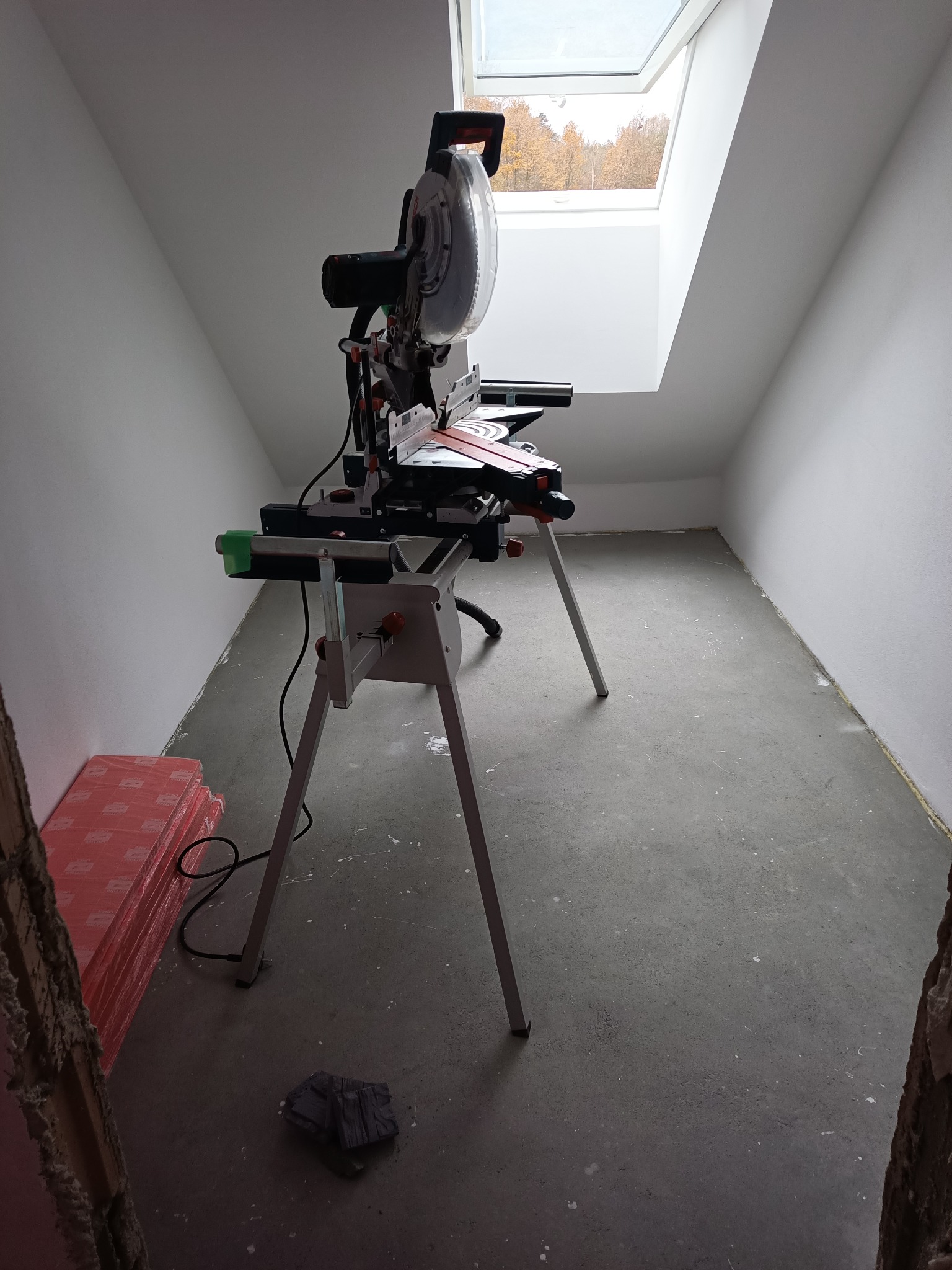 Ustawiona piła ukośnica na metalowym stojaku w surowym pokoju na poddaszu ze świetlikiem dachowym, obok leżą paczki paneli i szmatka.