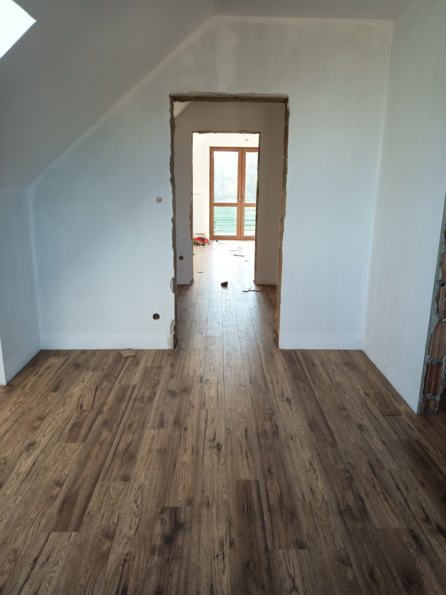 Wnętrze w trakcie remontu: świeżo położone panele podłogowe z efektem drewna, surowe ściany przygotowane do malowania, widoczne niedokończone ościeżnice drzwiowe.