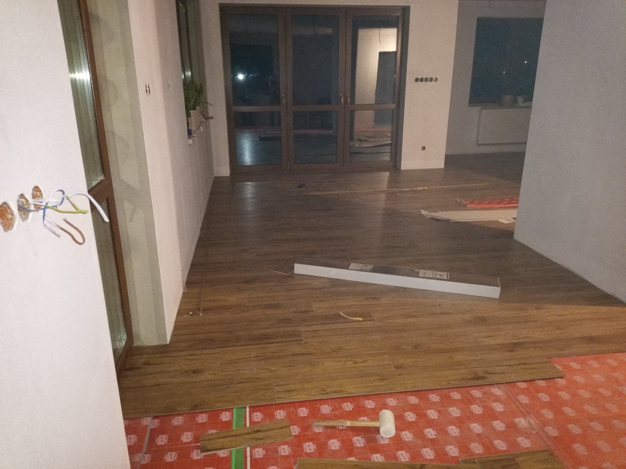 Wnętrze w trakcie wykańczania: brązowe panele podłogowe imitujące drewno, częściowo ułożone na czerwonej macie wygłuszającej, biały gumowy młotek, paczka paneli w tle, ściany w trakcie malowania...