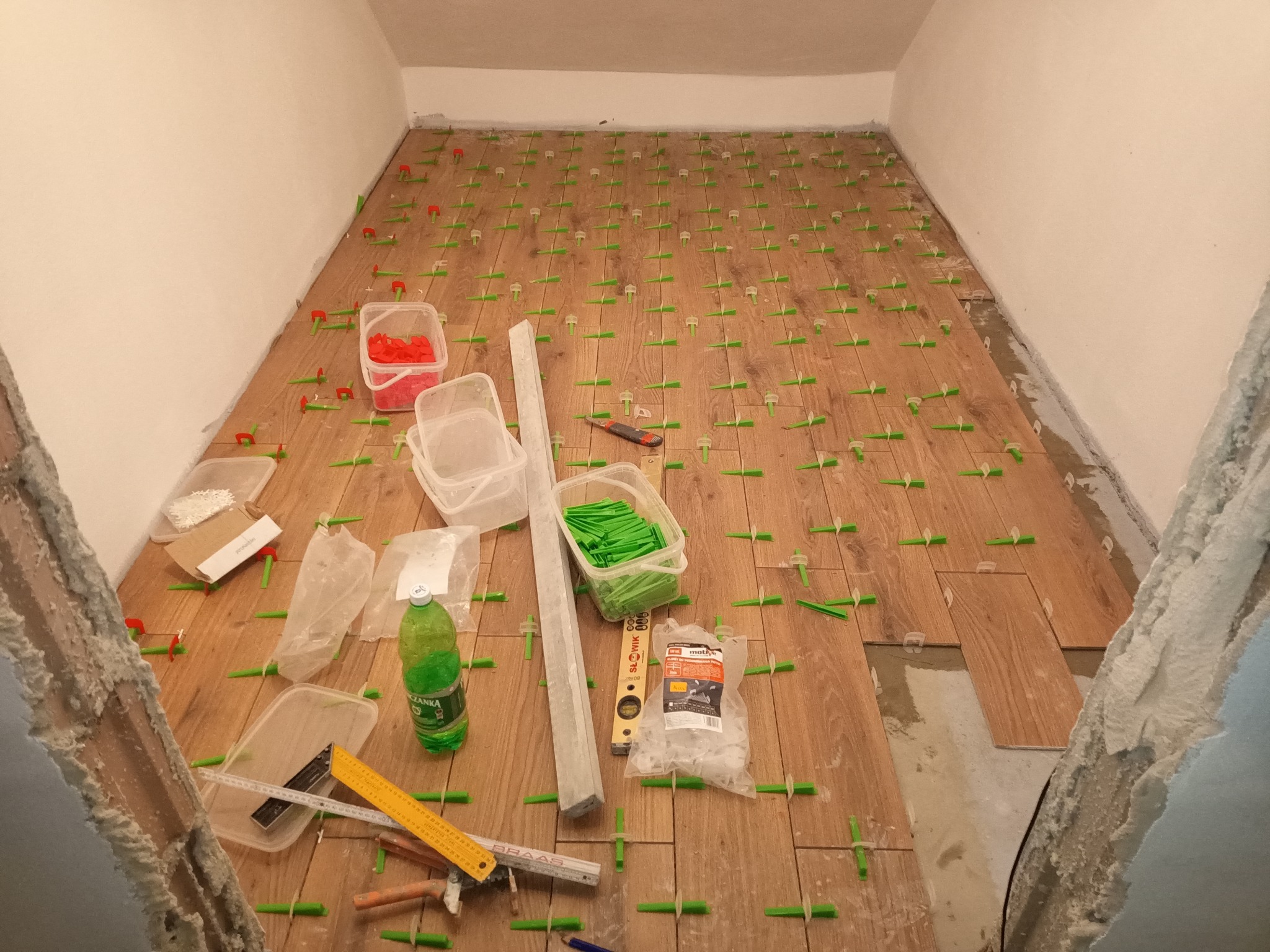Proces układania drewnopodobnych płytek podłogowych z użyciem zielonych krzyżyków dystansowych, poziomicy, miarki, nożyka i opakowań z zaprawą, w pomieszczeniu z surowymi ścianami.