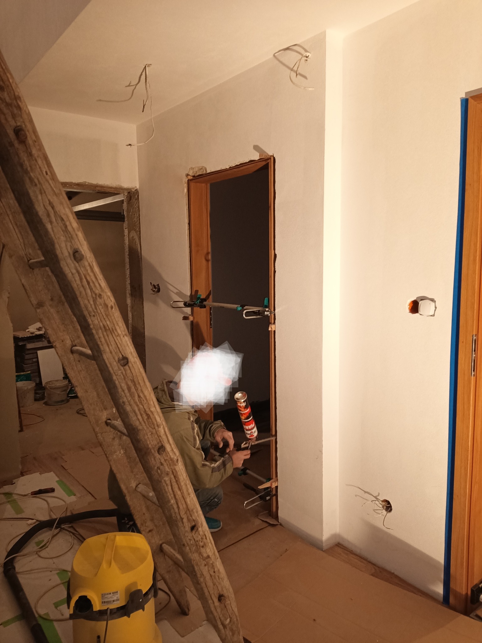 Montaż ościeżnicy w Tarnowie: fachowiec uszczelnia przestrzeń między ścianą a drewnianą ramą drzwi przy użyciu pistoletu do pianki montażowej, widoczna drabina i odkurzacz przemysłowy.