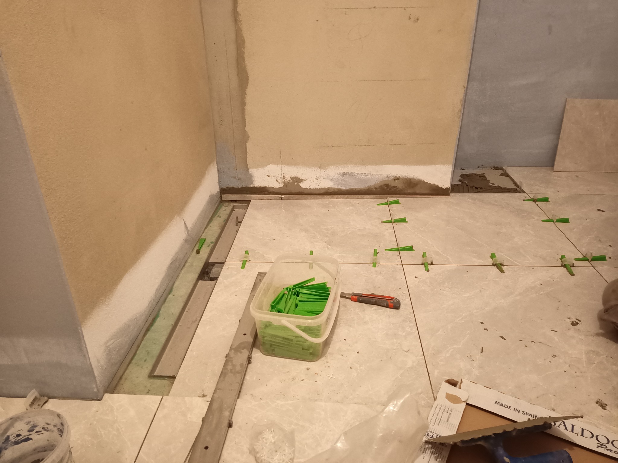 Fragment łazienki w trakcie prac wykończeniowych: ułożone płytki podłogowe z zielonymi krzyżykami dystansowymi, widoczny pojemnik z zapasem krzyżyków, nóż tapicerski i paczka płytek z napisem 'MADE...