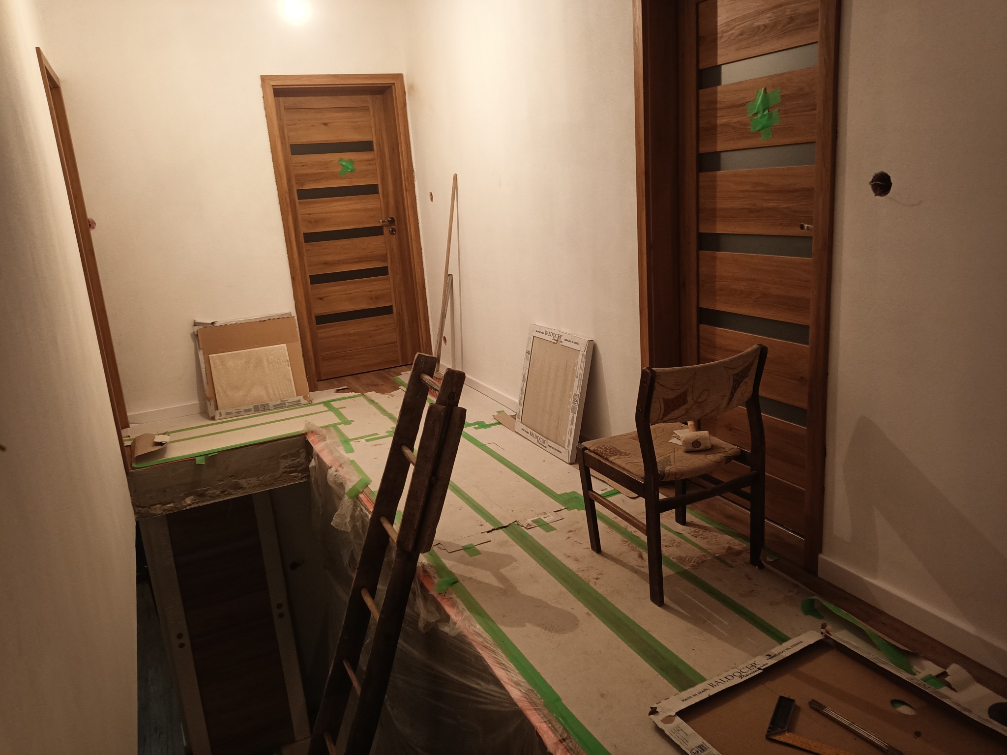 Wnętrze w trakcie prac wykończeniowych: widoczne zabezpieczone podłoże, drabina, krzesło, oraz zamontowane drewniane drzwi z zieloną taśmą zabezpieczającą, obok drzwi w trakcie montażu.