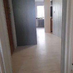 Widok na korytarz z wykończoną podłogą w odcieniach bieli, częściowo widoczne drzwi po lewej stronie i grzejnik w głębi