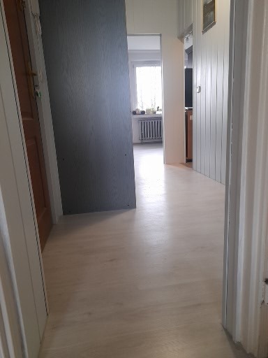 Widok na korytarz z wykończoną podłogą w odcieniach bieli, częściowo widoczne drzwi po lewej stronie i grzejnik w głębi