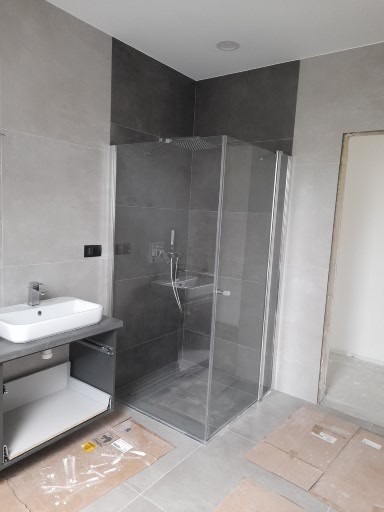 Nowoczesna łazienka w szarościach z kabiną prysznicową typu walk-in, umywalką nablatową i otwartą szafką z wysuwaną szufladą; na podłodze kartony zabezpieczające.