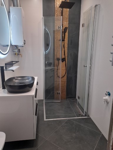 Nowoczesna łazienka z czarną umywalką na białej szafce, kabiną prysznicową typu walk-in z deszczownicą i grzejnikiem gazowym z oświetlonym lustrem.