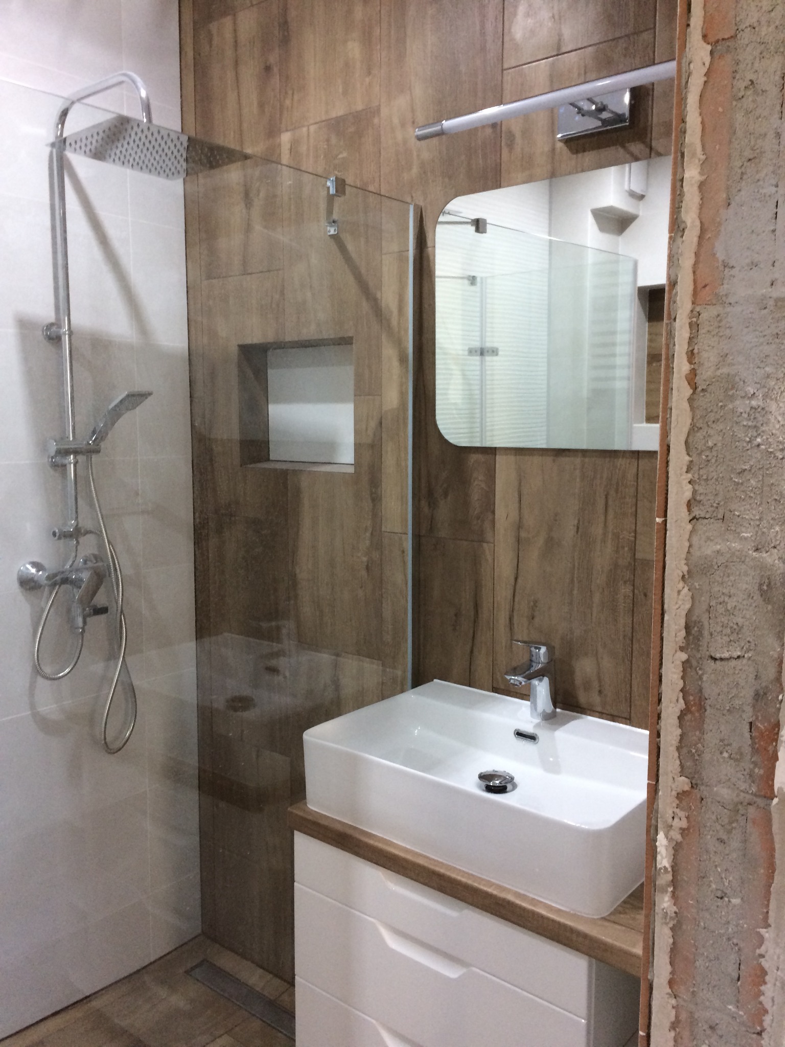 Nowoczesna łazienka z kabiną prysznicową typu walk-in, umywalką nablatową i drewnopodobnymi płytkami ściennymi, widoczny fragment ściany w trakcie prac wykończeniowych.