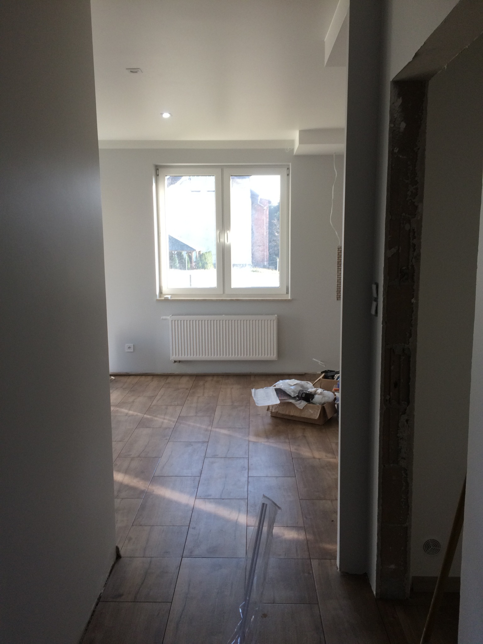 Wnętrze pokoju w Drogomyślu w trakcie wykańczania, widoczne płytki podłogowe imitujące drewno, grzejnik pod oknem i kartony z materiałami budowlanymi.