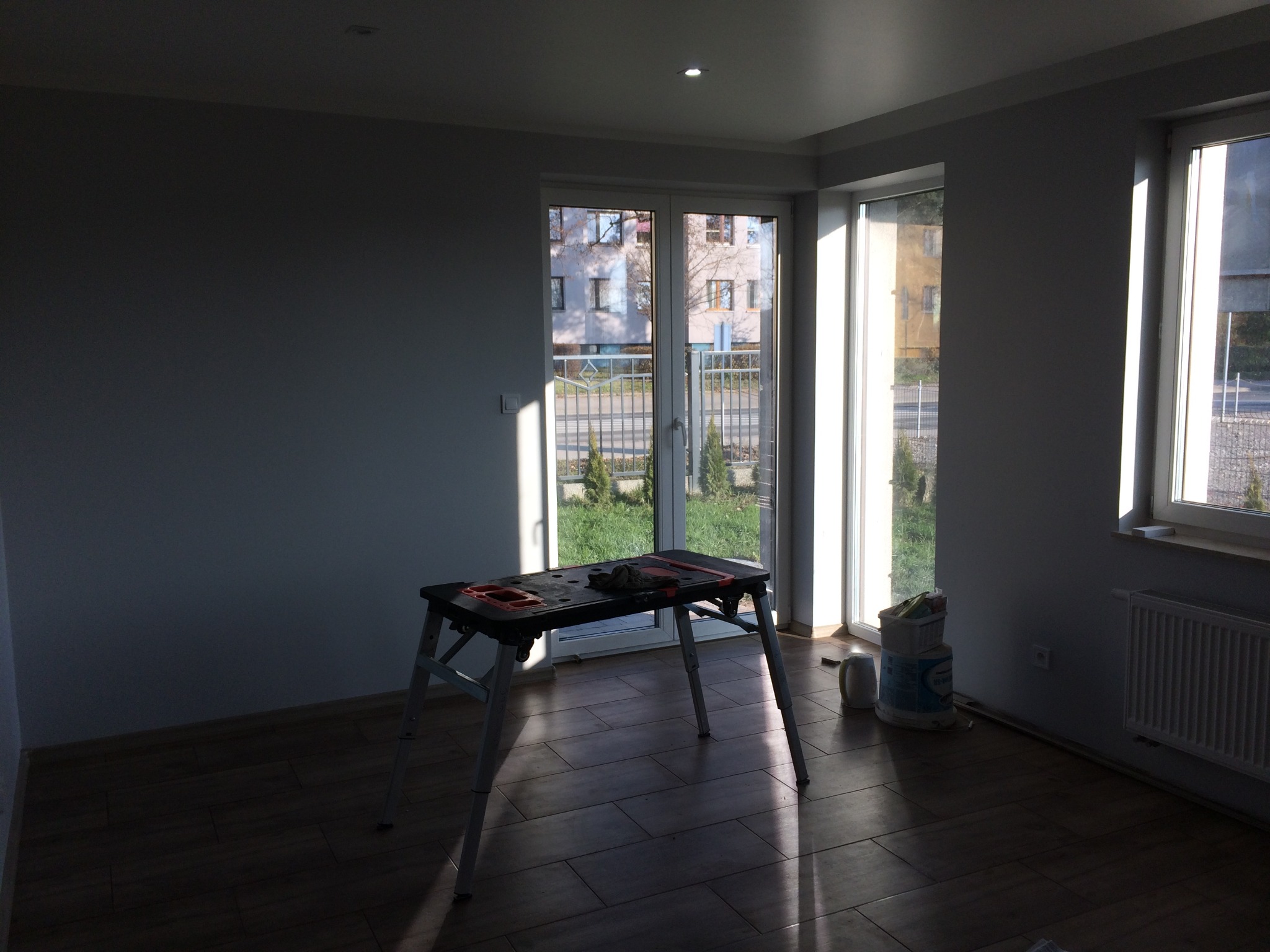 Przygotowania do malowania ścian w łazience: rozstawiony stół roboczy, wiadro z farbą, widoczne okno i drzwi balkonowe, podłoga wyłożona panelami.