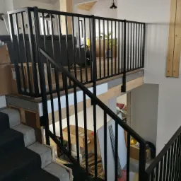 Czarne, metalowe poręcze schodowe i balustrada na antresoli w nowoczesnym wnętrzu, widok z góry na dół, częściowo widoczne kartony i inne przedmioty.