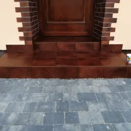 Wejście do domu z brązowymi schodami wyłożonymi płytkami, ceglana obudowa drzwi, szara kostka brukowa na podwórku, kubek z kwiatowym wzorem na schodach.